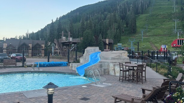 A pool area with a blue slide, outdoor seating, and a view of a forested hill.