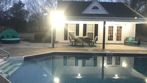 Una piscina con un área de patio, una zona de estar techada y una casa con porche.