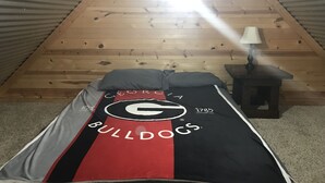 Un dormitorio triangular en el ático, con techo inclinado, una cama individual con manta temática de Bulldog, una mesita de noche con lámpara y un tragaluz visible.