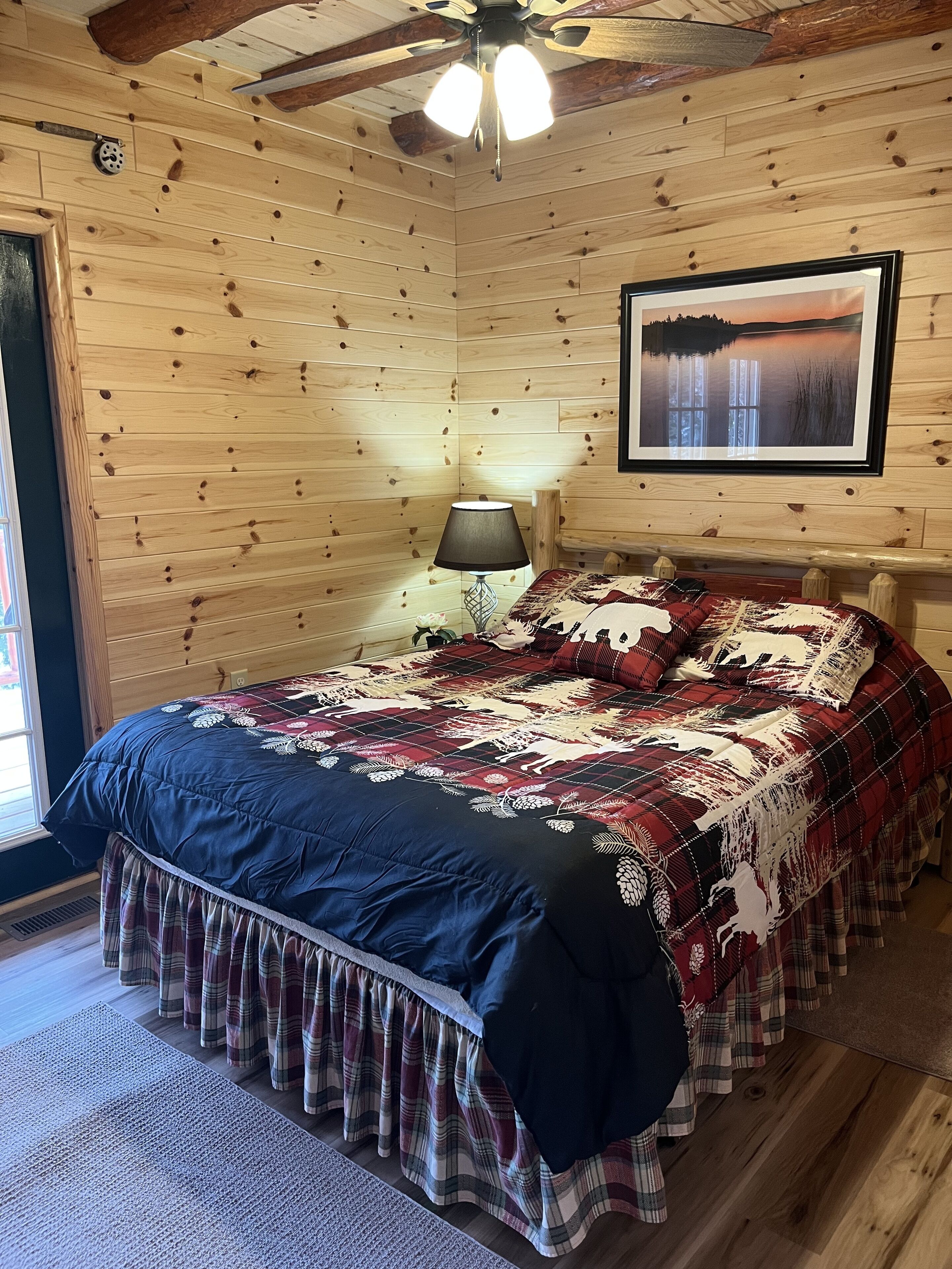 A wooden cabin bedroom with a bed, a ceiling fan, a lamp, and a framed picture on the wall.