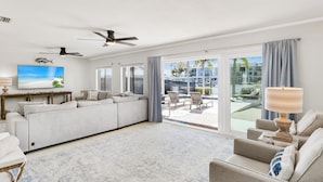 A spacious living room with a large sofa, a coffee table, and a TV mounted on the wall. There are ceiling fans, a large window with a view of the pool and outdoor seating, and a lamp on the side table.
