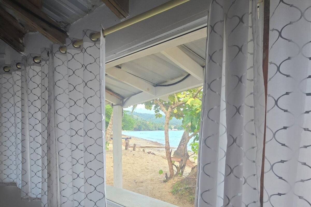Ein Zimmer mit großem Fenster, weißen Vorhängen mit Muster und Blick auf Strand und Palmen.