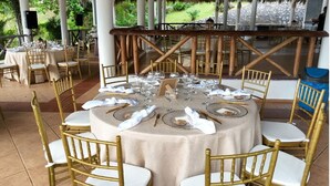 Een overdekt terras met ronde tafels gedekt voor een maaltijd, omringd door gouden stoelen.