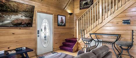 Interior de una cabaña de madera con una escalera, un sofá, una mesa y un televisor que muestra una escena de la naturaleza.