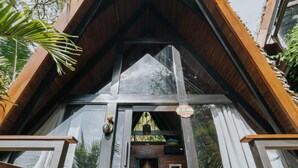 Une cabane moderne, dotée d’un toit triangulaire, de grandes fenêtres en verre, d’une terrasse en bois et d’une chambre à coucher visible, avec un lit et une table de chevet.