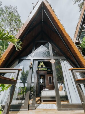 A modern, triangular-roofed cabin with large glass windows, a wooden deck, and a visible bedroom with a bed and nightstand. - Bohemia Experience (Cóbano)
