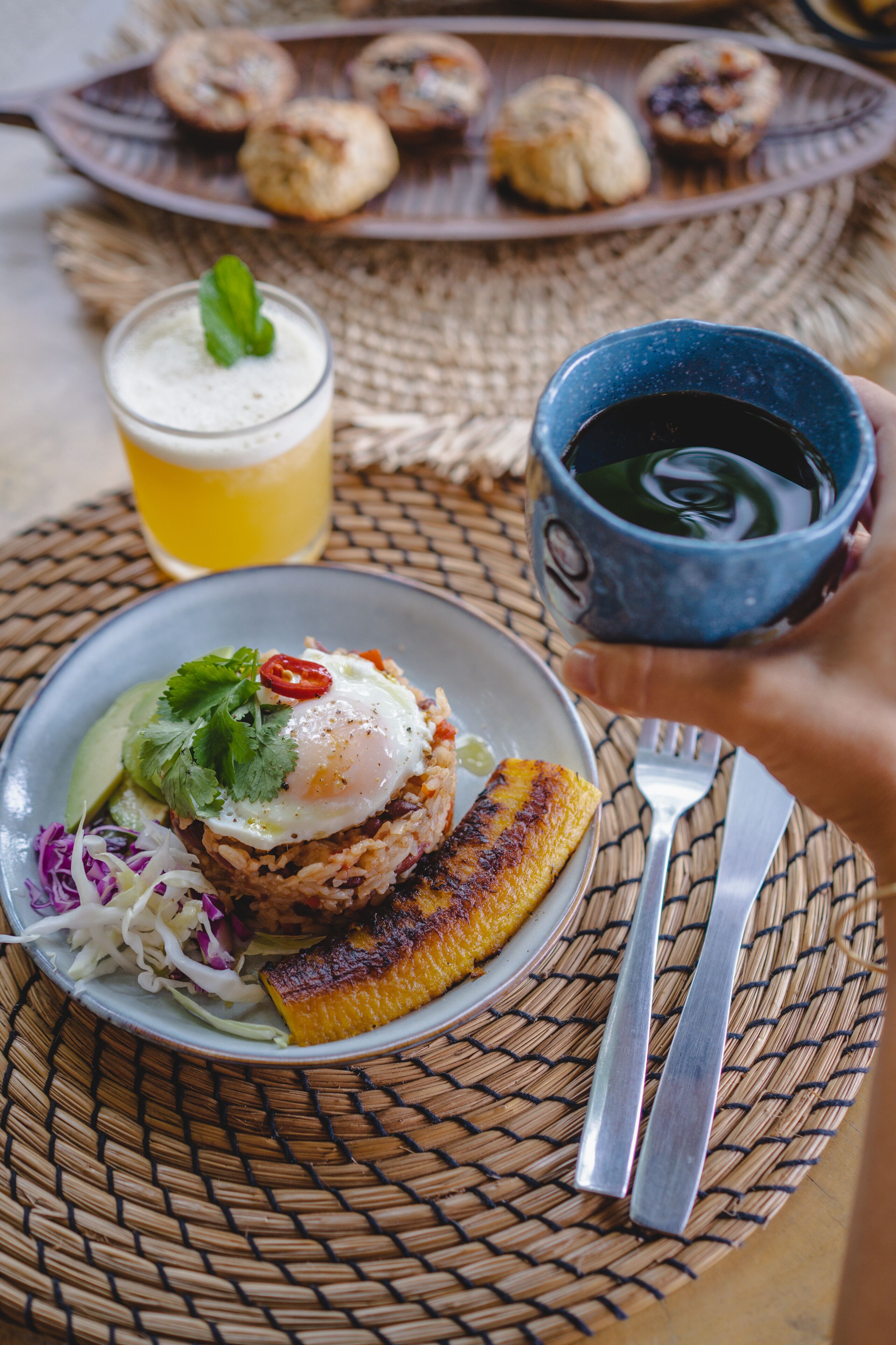 A plate of food with a fried egg, avocado, and red cabbage, a glass of beer, and a cup of coffee on a woven mat.