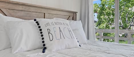 A bed with white bedding and a pillow with the text 'LIFE IS BETTER AT THE BEACH'.