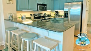 A kitchen with teal cabinets, a stainless steel refrigerator, a microwave, and a stove. There are three bar stools and a small island with a sink. - Lazy River and Indoor Pool Sleeps 8 CT605 (Gulf Shores)