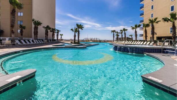 A large outdoor swimming pool with multiple lanes, surrounded by palm trees and lounge chairs, with high-rise buildings in the background. - Lazy River and Indoor Pool Sleeps 8 CT605 (Gulf Shores)