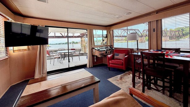 Ein Zimmer mit Fernseher, Schreibtisch, Stuhl, Tisch und großen Fenstern mit Blick auf eine Brücke und Wasser.