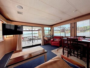 A room with a television, a desk, a chair, a table, and large windows with a view of a bridge and water.