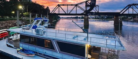 A houseboat docked at a pier with a metal bridge overhead, a calm river, and a sunset sky.