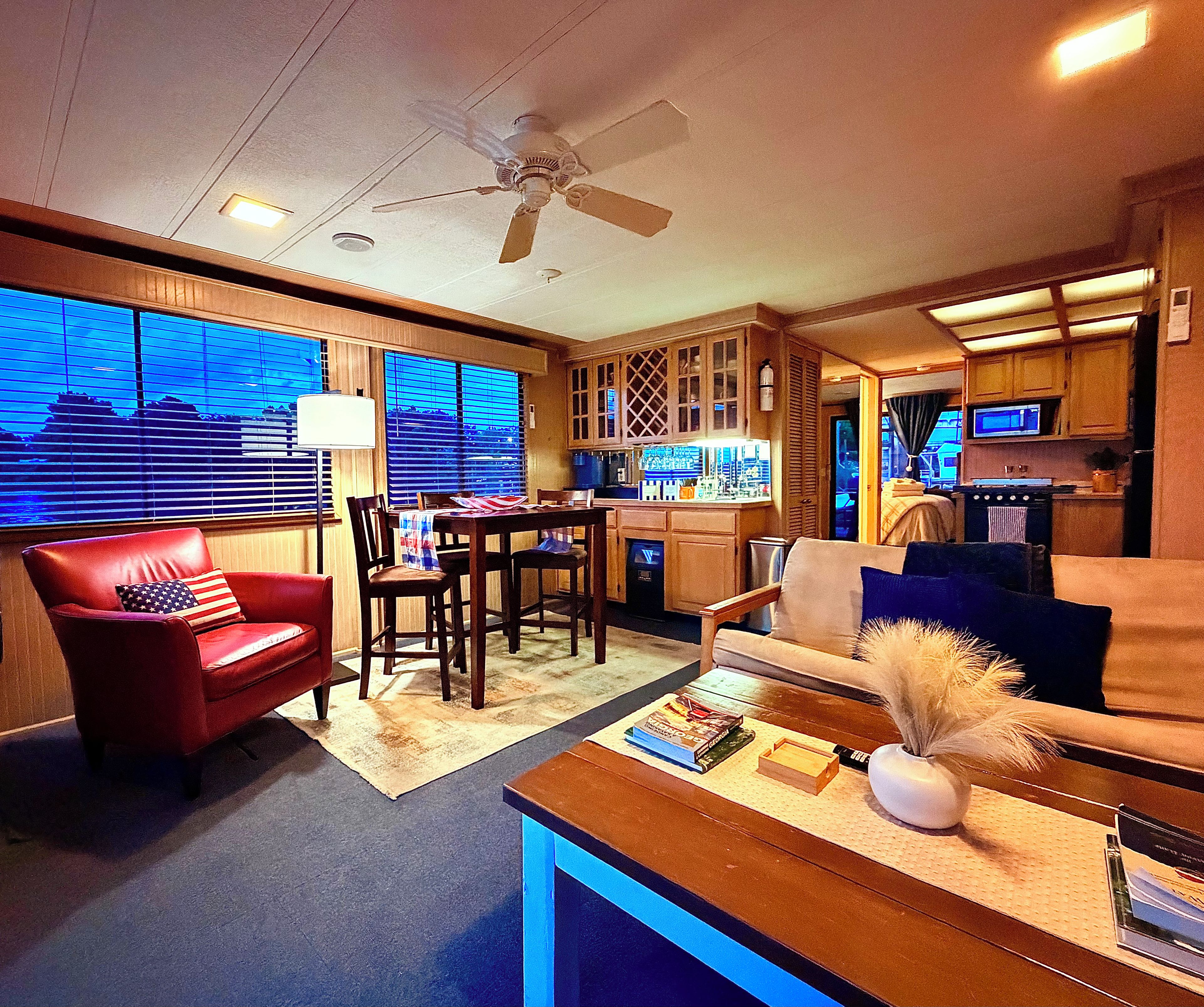 A compact living space with a dining area, a red armchair, a sofa, a ceiling fan, and a view through large windows.