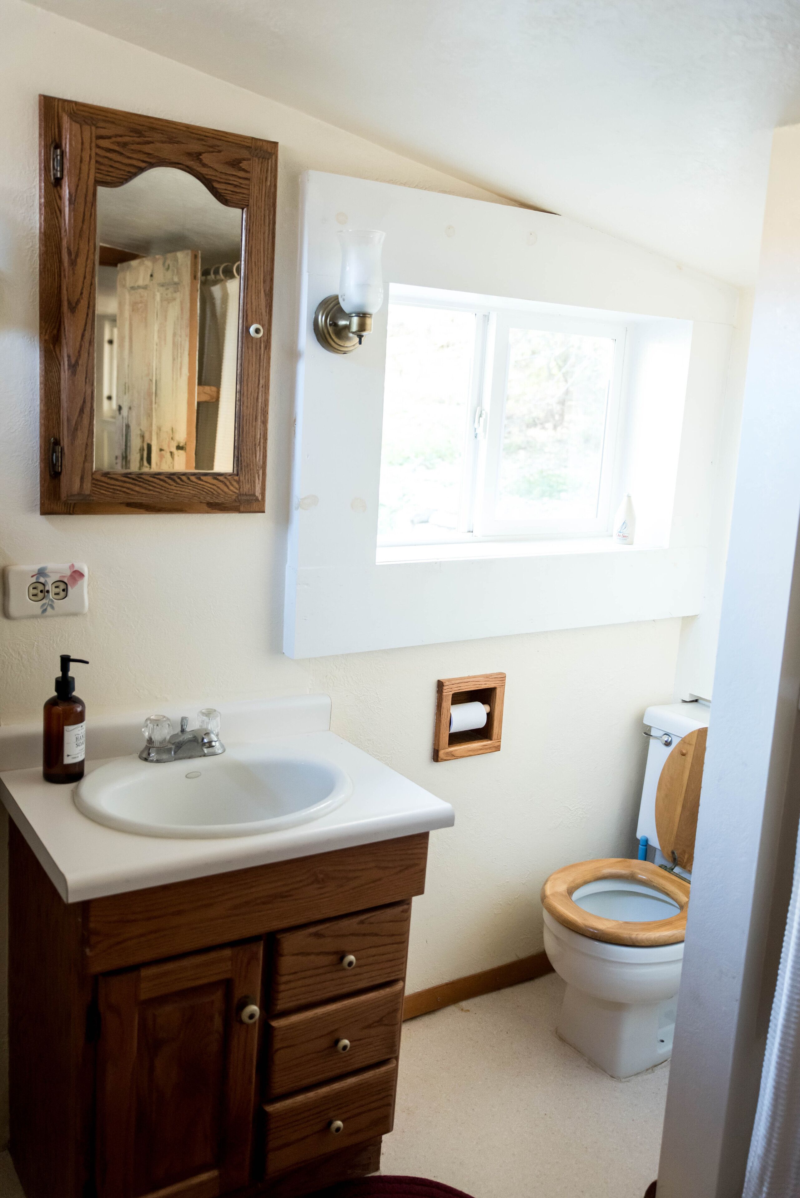 A bathroom with a wooden vanity, a white sink, a mirror, a window, and a toilet.