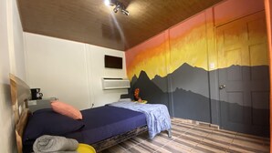 Ett sovrum med en säng, ett litet bord med handdukar, en kaffemaskin, en luftkonditioneringsanläggning och en vägg med en målning av ett berg.