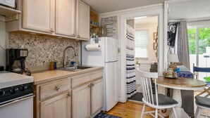 A compact kitchen with a stone backsplash, a built-in stove, a refrigerator, and a dining area with a chair and table. - Cozy Mid-Cape Studio Steps to the Beach & Pool (Dennis Port)