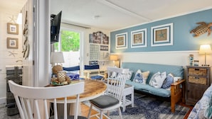 A cozy living room with a blue wall, a wooden dining table, a white sofa, and a TV mounted on the wall. - Cozy Mid-Cape Studio Steps to the Beach & Pool (Dennis Port)