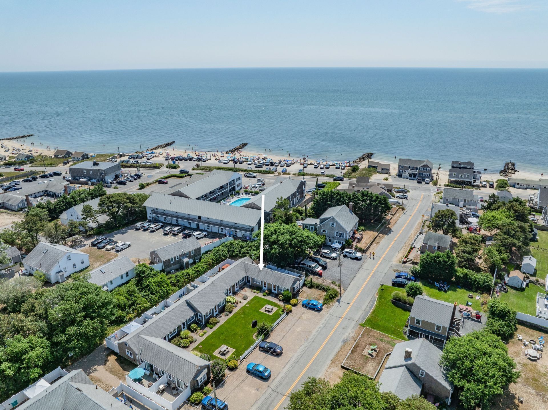 An aerial view of a coastal town with a prominent building featuring a swimming pool, surrounded by residential houses and a beach area.