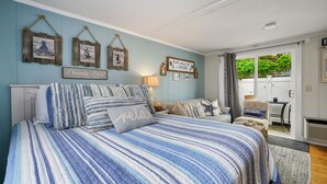 A bedroom with a bed, a nightstand, a lamp, and a view of the outdoors. - Charming Dennis Port Getaway for Two with Pool & Walk to Beach (Dennis Port)