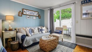 A cozy bedroom with a bed, bedside table, lamp, and a view of the outdoors through a sliding door. - Charming Dennis Port Getaway for Two with Pool & Walk to Beach (Dennis Port)
