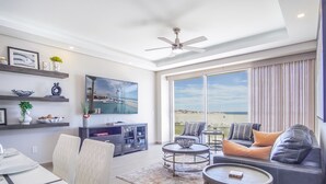 Un vaste espace de vie doté d’une grande fenêtre donnant sur la plage, d’un ventilateur de plafond et d’un coin repas avec une table et des chaises.