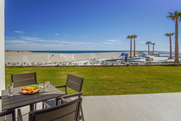 A patio with a table set for two, overlooking a pool area with palm trees and a view of the beach. - Spectacular 2 bedroom condominium Encantame E102 (Puerto Peñasco)