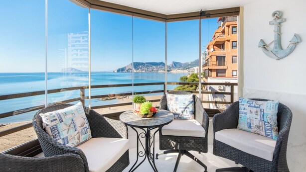 Un balcón con muebles de mimbre, una pequeña mesa con fruta y vistas al mar y edificios.