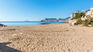 Una playa arenosa con cielo despejado, un promontorio rocoso y edificios al fondo.