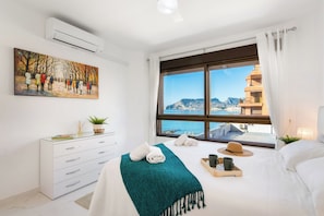 A hotel room with a bed, a window with a view of the sea and buildings, a painting on the wall, and a white dresser.