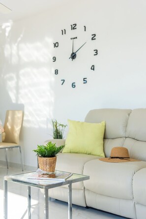 A living room with a sofa, a glass coffee table, a wall clock, a potted plant, and a hat on the sofa.