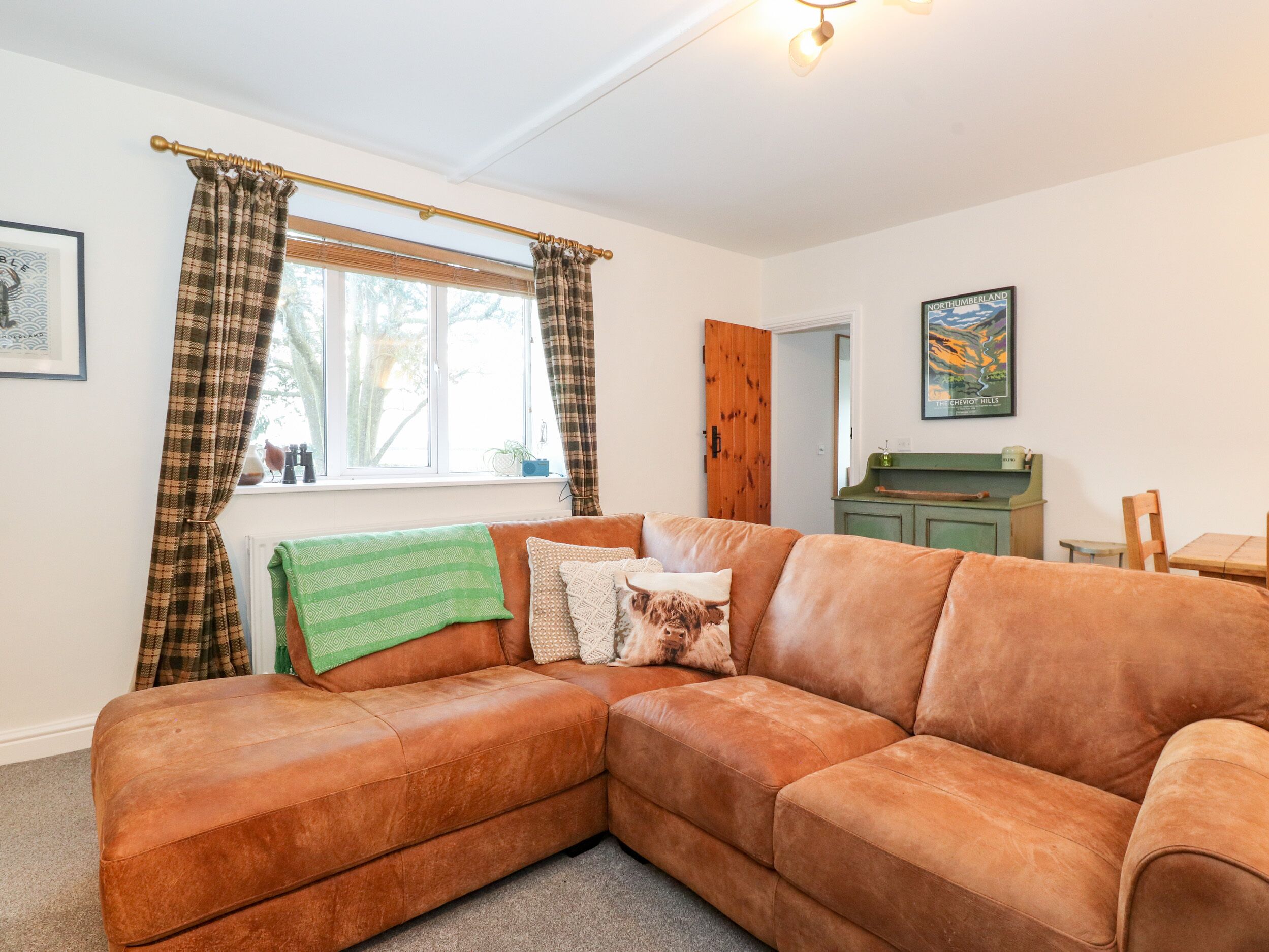A cozy living room with a brown leather sectional sofa, a green throw blanket, and plaid curtains.