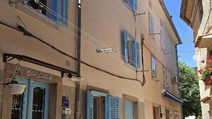 A narrow street with buildings featuring blue shutters and doors, a small outdoor seating area, and a clear sky. - Apartments Blue Lavender (Cres)