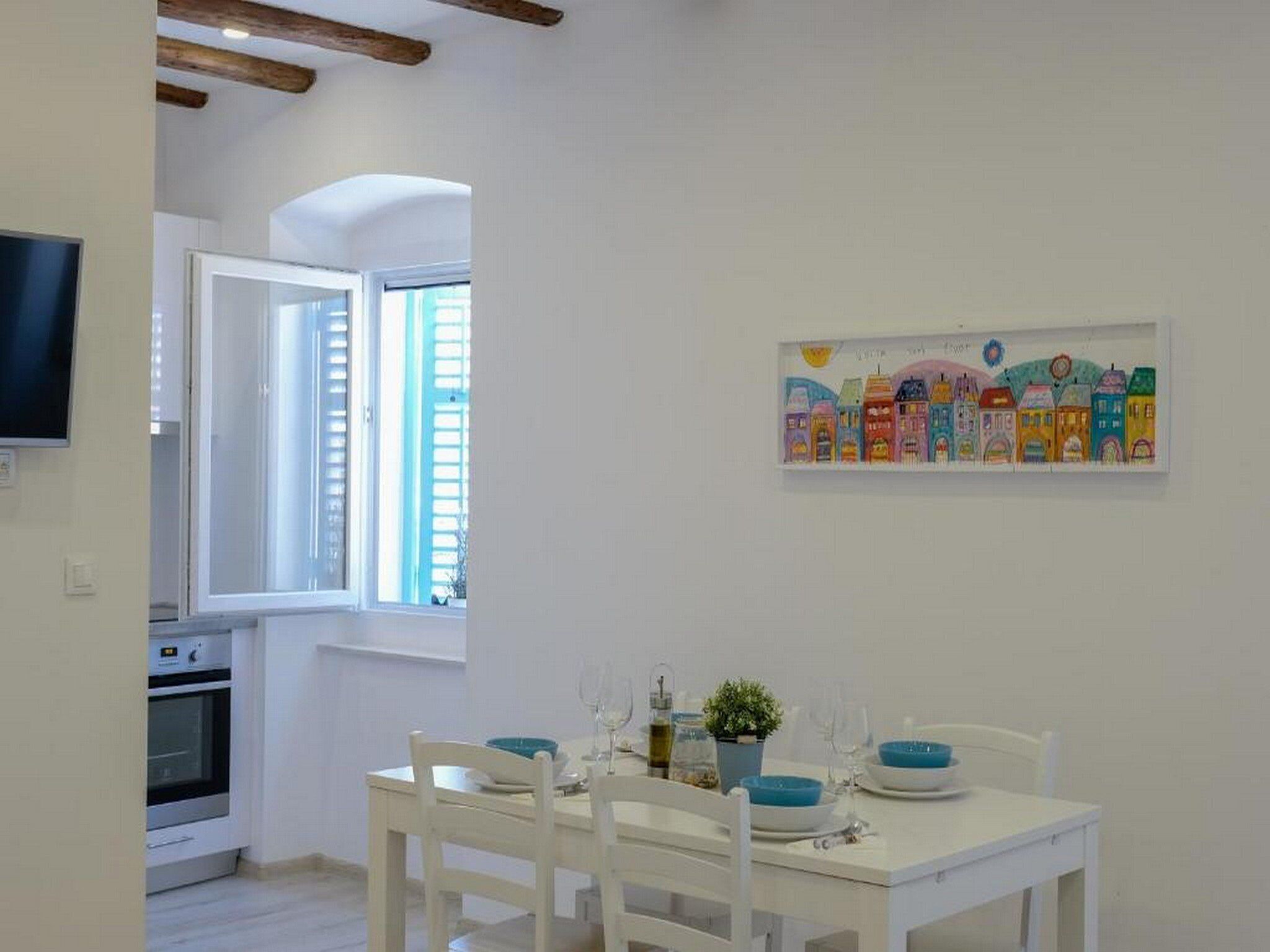 Ein Essbereich mit einem weißen Tisch, gedeckt für zwei Personen, einem Fenster mit Jalousien und einem farbenfrohen Bild an der Wand.