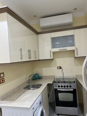 A compact kitchen with white cabinets, a built-in washing machine, a gas stove, and a wall-mounted air conditioner. - Captivating 1-bed Apartment in Egypt (Shurtah al-Dakhlah)