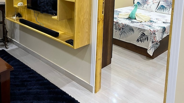 Quarto com um suporte de televisão de madeira, cama com colcha floral e tapete azul escuro.