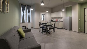 A modern apartment with a grey sofa, dining table, and chairs, a kitchen area with a refrigerator, and a window with curtains. - San Luca Apartments - Grillo by Wonderful Italy (Genoa)