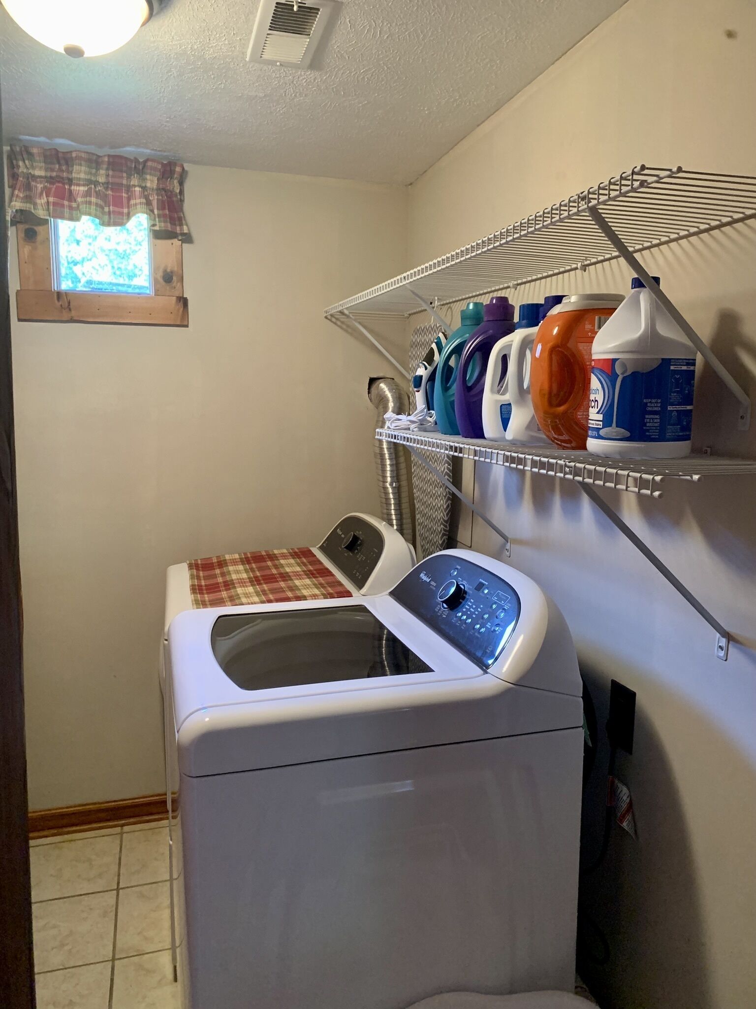 Une buanderie équipée d’un lave-linge à chargement frontal, d’un étendoir avec différents produits de lessive, et d’une fenêtre agrémentée d’un rideau à carreaux.