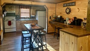 A rustic kitchen with wooden cabinets, a stove, and a dining area with stools. A sign on the wall reads "DO NOT FEED THE BEARS".