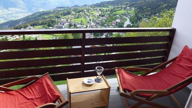 A balcony with two wooden chairs and a small table, overlooking a scenic valley with houses and a winding road. - Nice apartment for 4 people with WIFI, TV, balcony and panoramic view (Bluche)