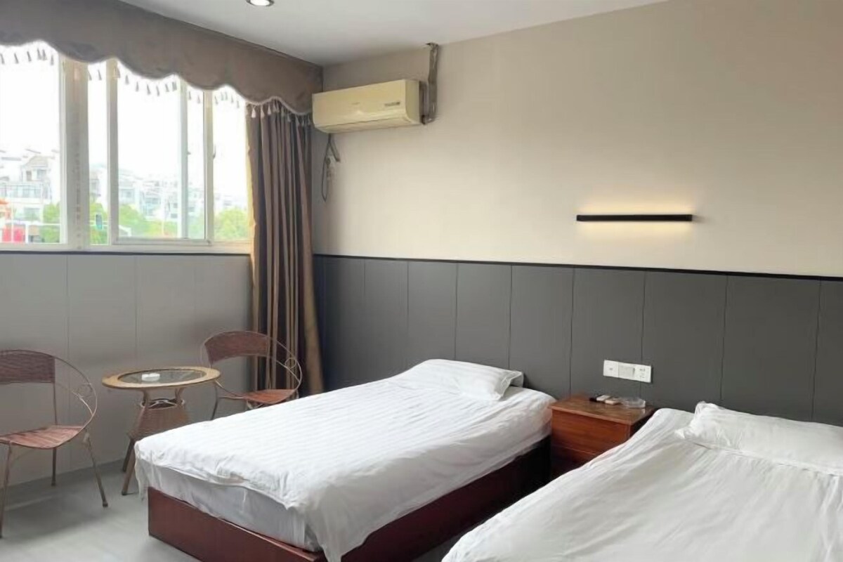 Habitación de hotel con dos camas, una mesita, dos sillas, un ventanal con cortinas, un equipo de aire acondicionado y una lámpara fijada a la pared.