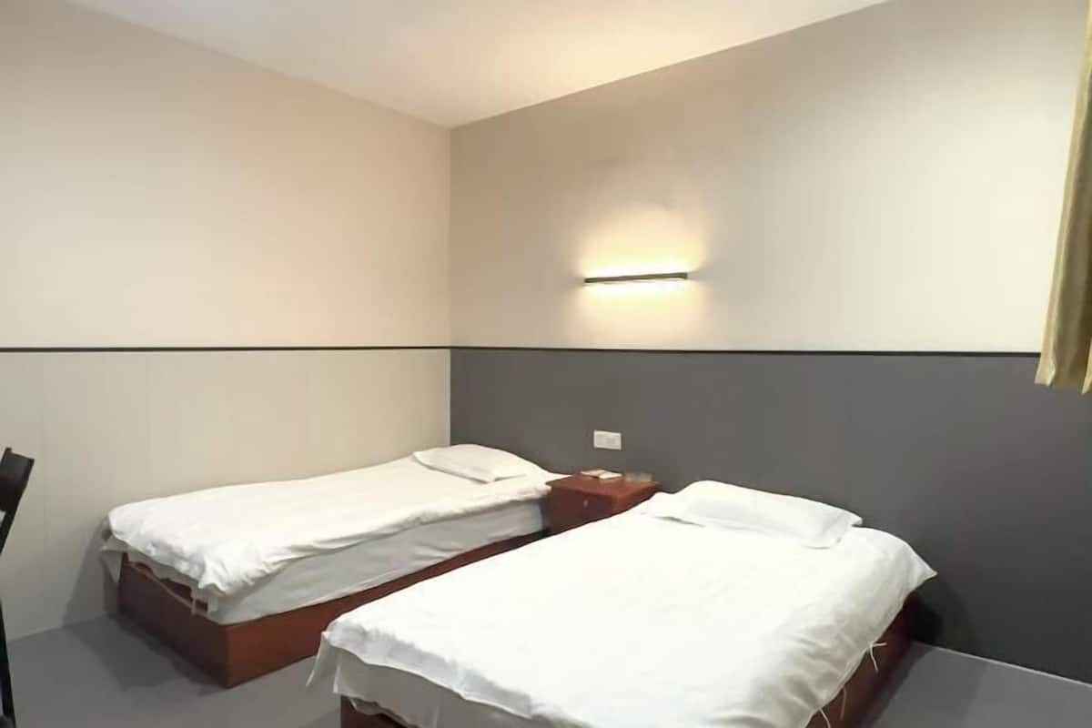 A hotel room with two single beds, a nightstand, a wall-mounted light, and a plain wall.