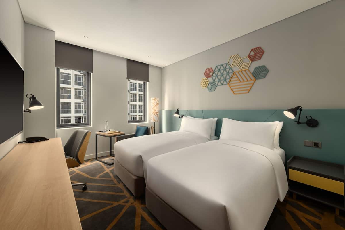 A hotel room with two beds, a desk, a chair, and a small table. There are windows and a wall decoration.
