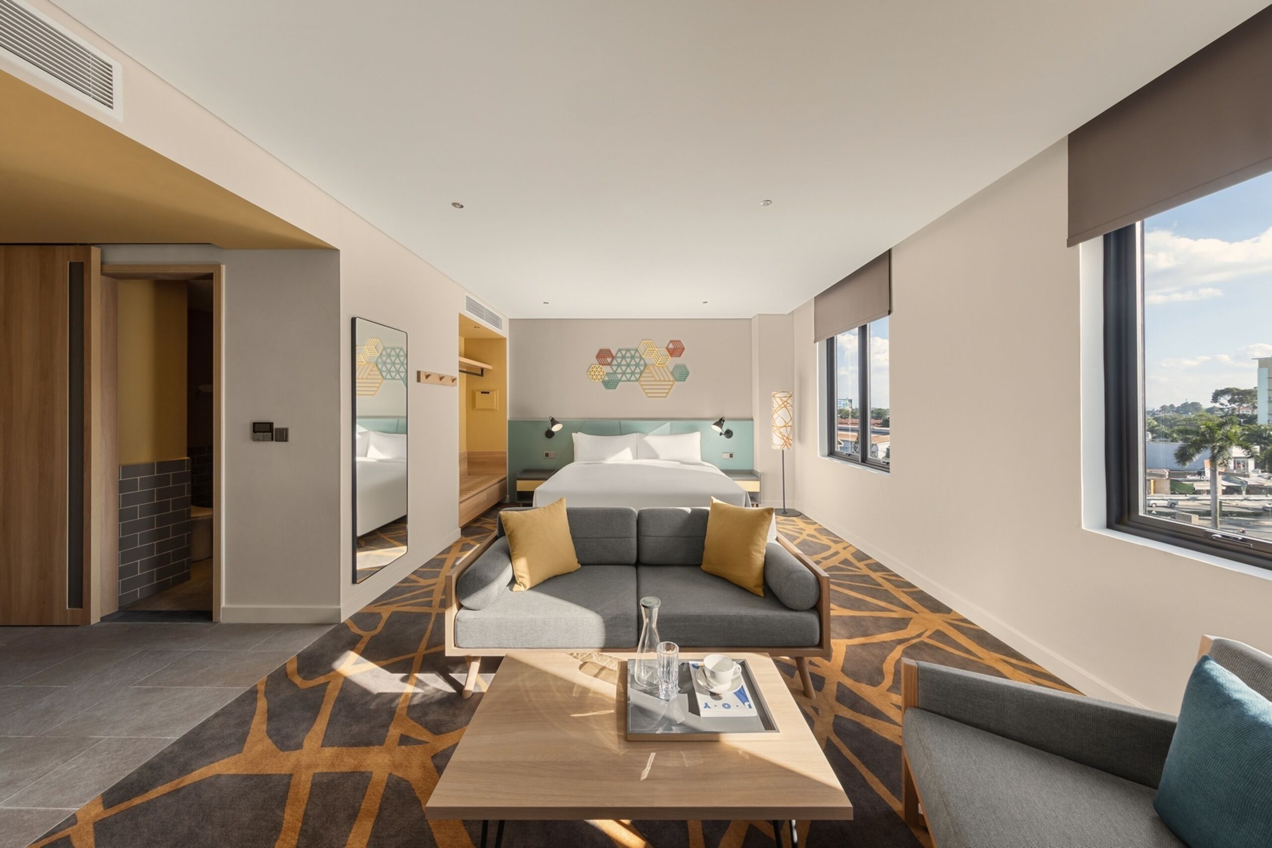 A modern hotel room with a large bed, a sofa, a dining table, and a view of the city.