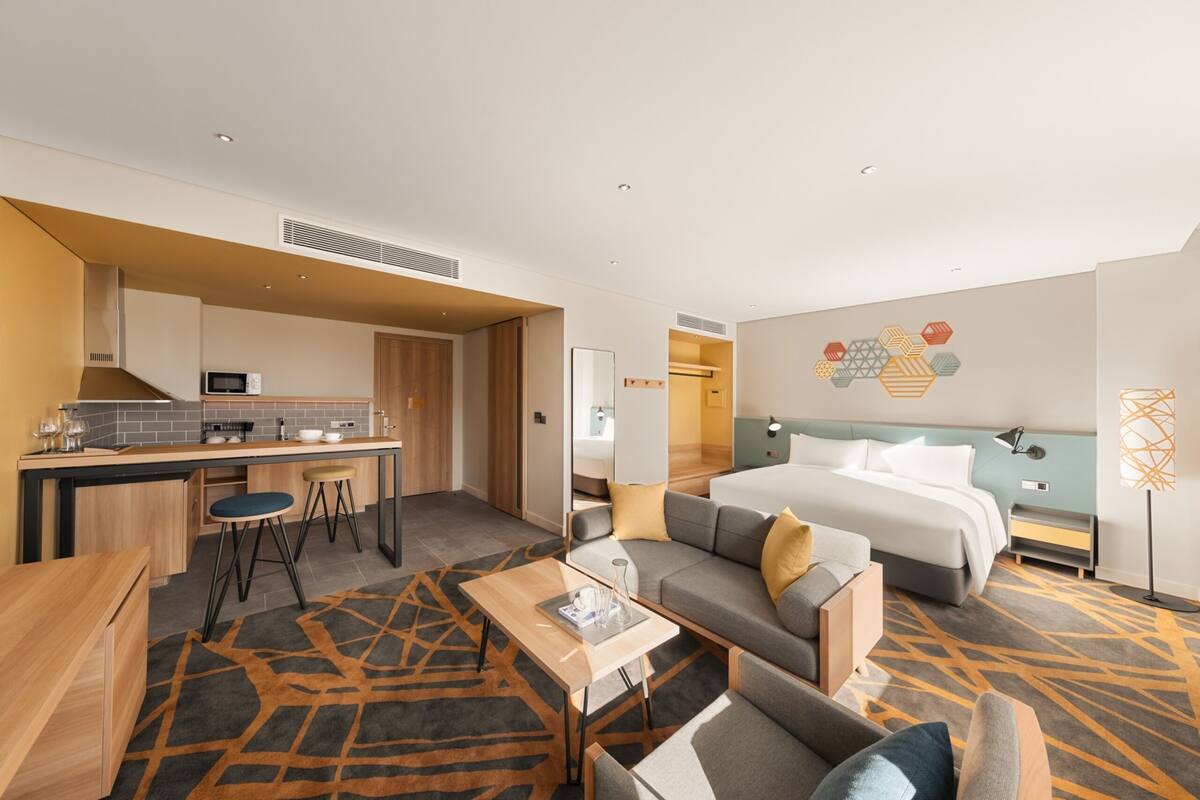 A modern hotel room with a kitchenette, a sofa, a bed, and a dining area.