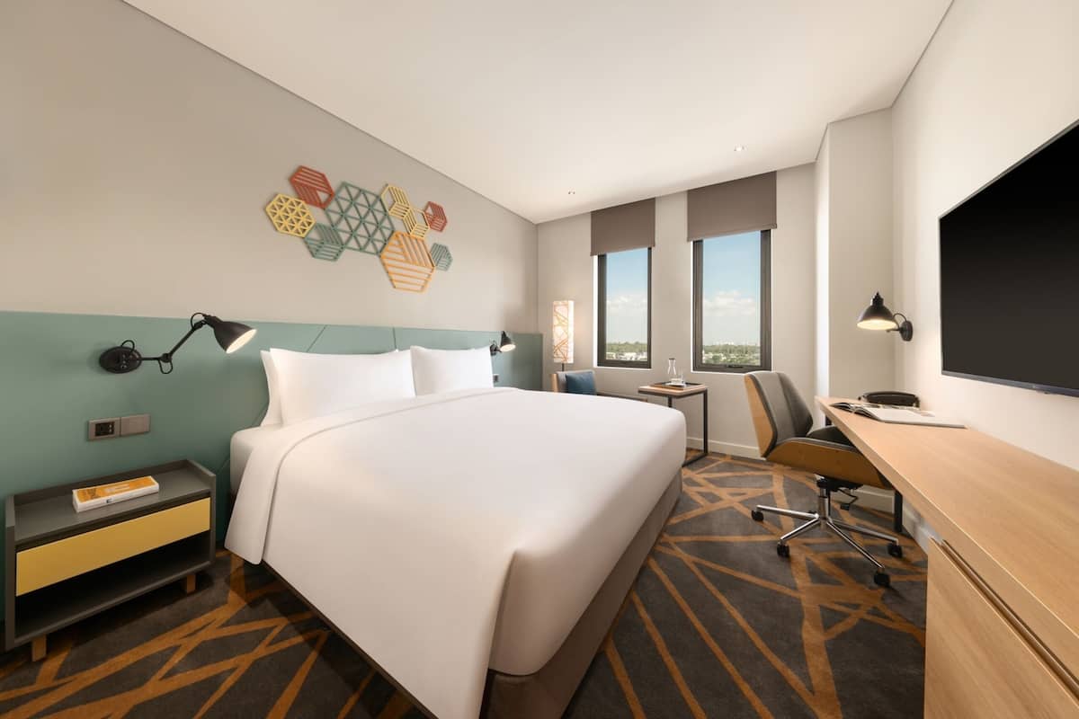 A hotel room with a large bed, a desk, a chair, a TV, and a window with a city view.
