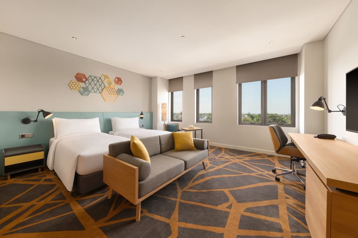A hotel room with two beds, a sofa, a desk, and a large window offering a city view.