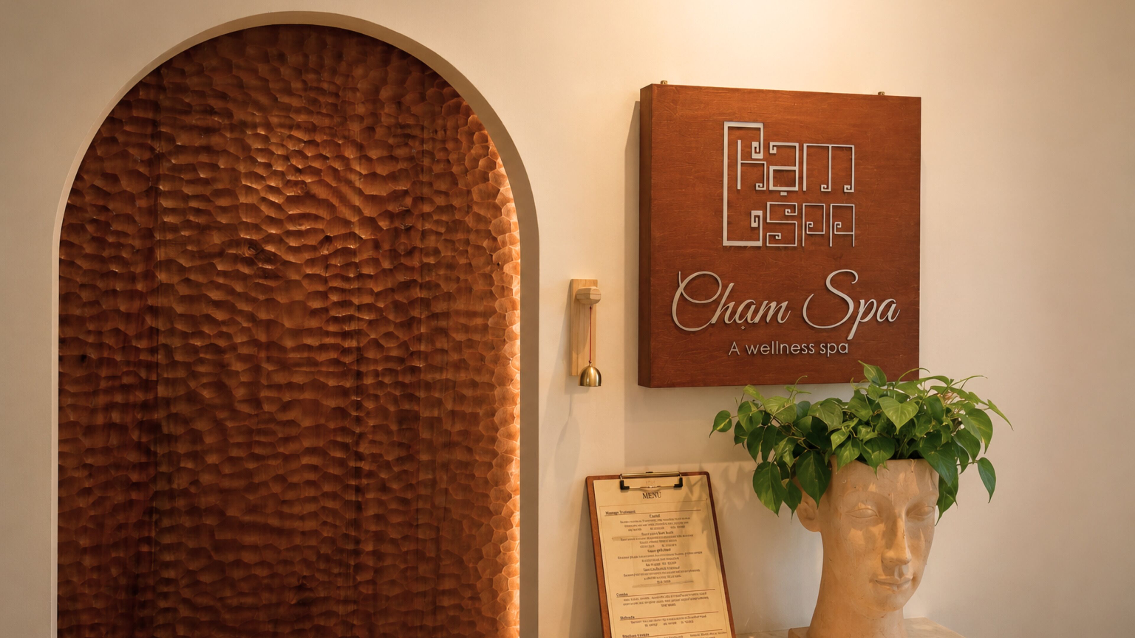 A spa entrance with a textured wooden door, a sign reading 'Cham Spa,' a menu board, and a potted plant.