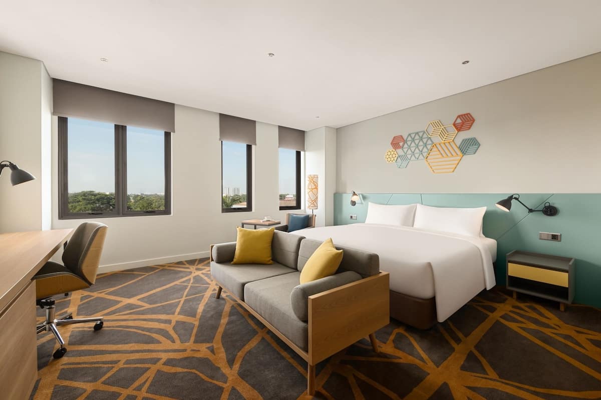 A modern hotel room with a large bed, a sofa, a desk, and a view of the city.