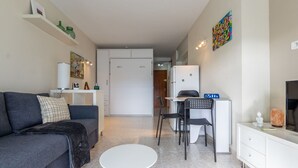 Un espace de vie compact comprenant un canapé, une petite table ronde, un bureau avec deux chaises, une armoire blanche et un tableau coloré accroché au mur.